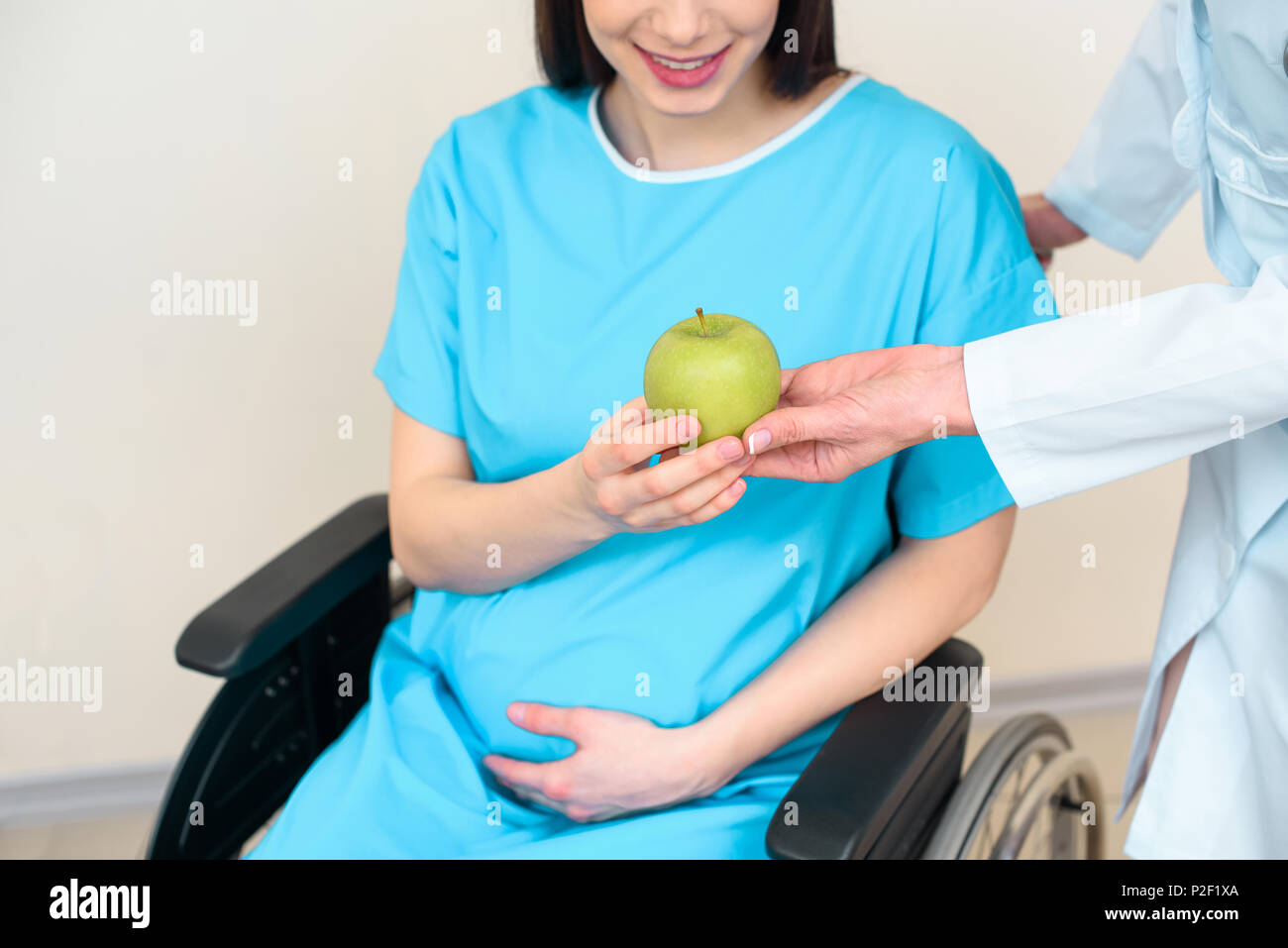 cropped shot of obstetrician gynecologist giving green apple to pregnant woman on wheelchair Stock Photo