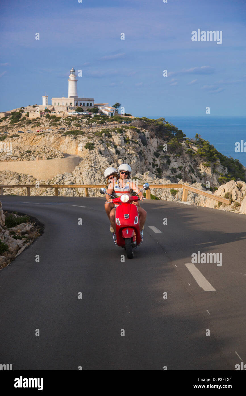 Young couple riding a red Vespa scooter on a coastal road along the Cap de Formentor peninsula with Faro de Formentor lighthouse Stock Photo