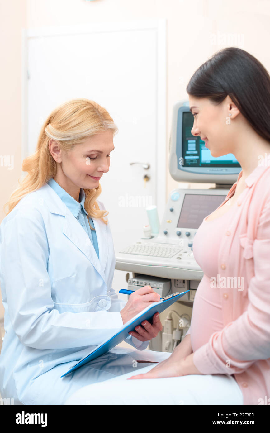 obstetrician gynecologist consulting happy young pregnant woman and writing in clipboard Stock Photo