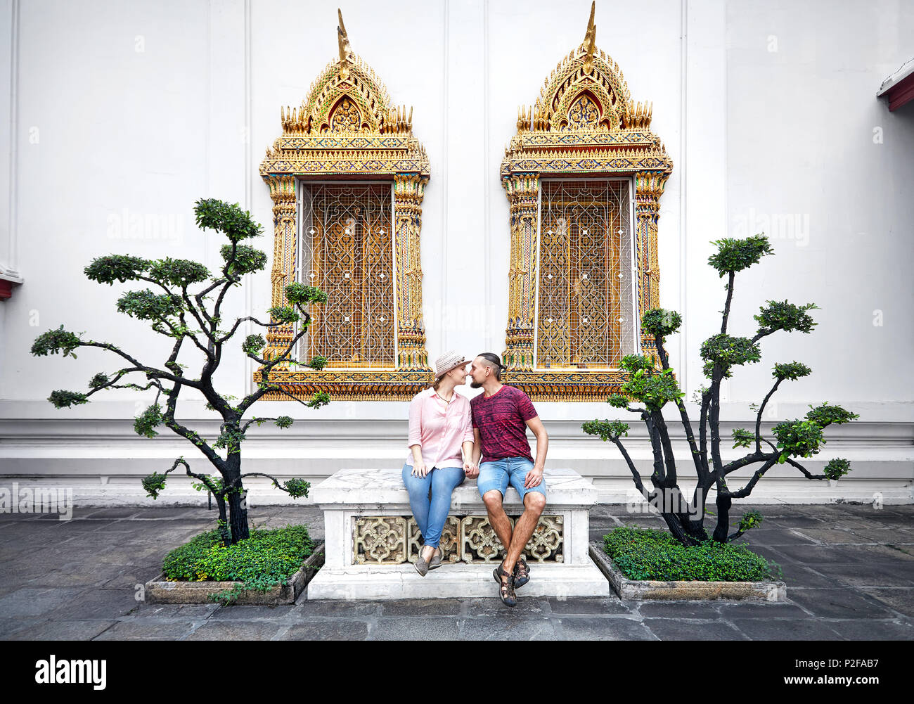 Young Couple in red clothes sitting on the bench near decorative trees and golden windows of Wat Pho in Bangkok, Thailand Stock Photo