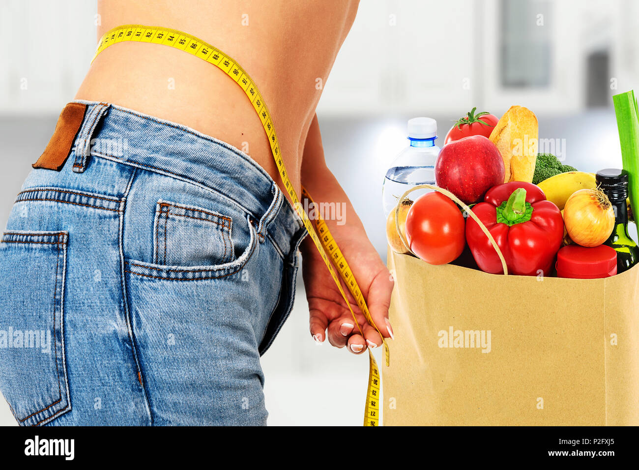 Healthy eating concept - woman in blue jeans measures her perfect waistline with a measuring tape next to a bag of dietetic food in the kitchen (mixed Stock Photo