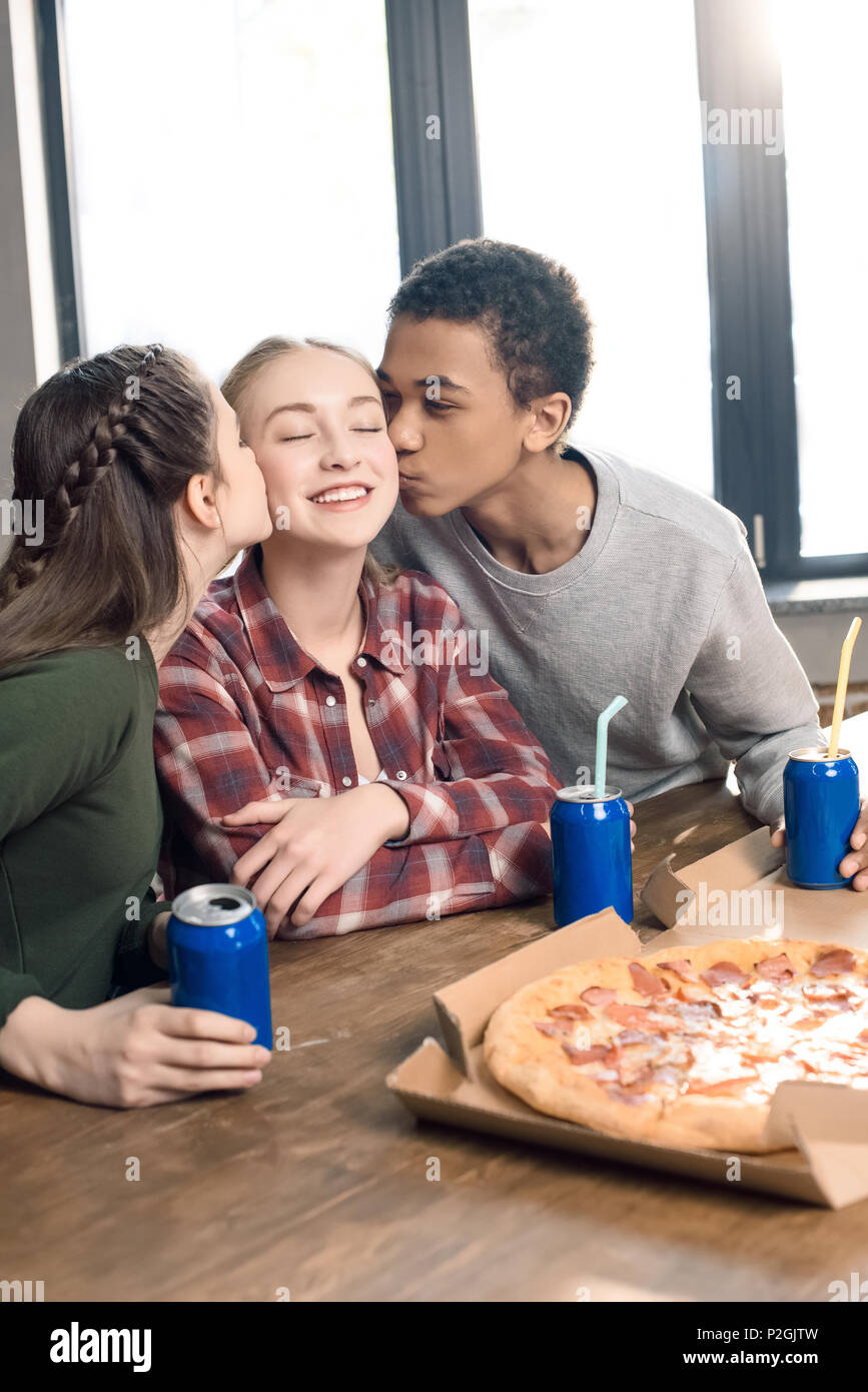 side view of friends kissing smiling teenage girl's cheeks, eating pizza at home concept Stock Photo