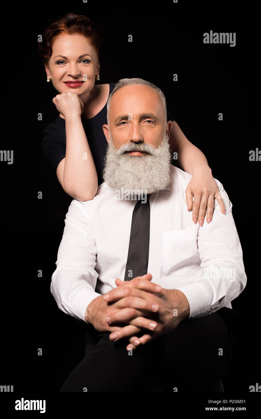 Half Length Shot Of Man Sitting On A Chair With His Wife Standing Behind And Leaning On His 