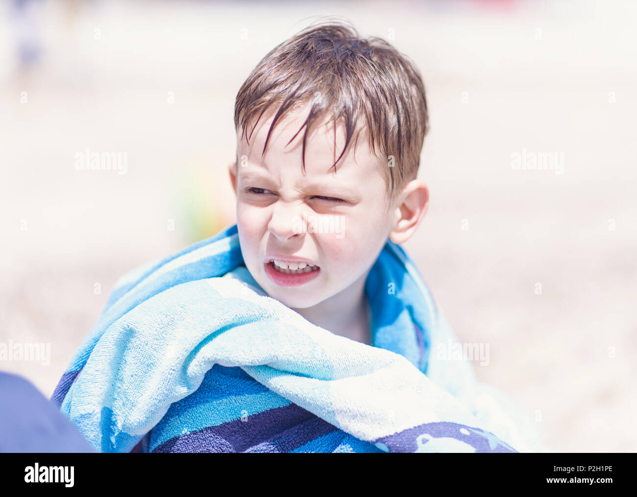 Wet young boy sitting on the beach with towel around his neck getting some heat in the sun Stock Photo