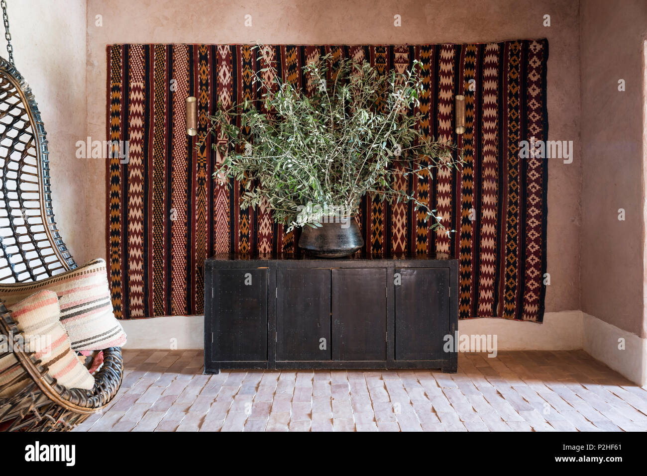 Woven hanging basket chair in room with earth walls, terracotta floor and kilim wall hang Stock Photo