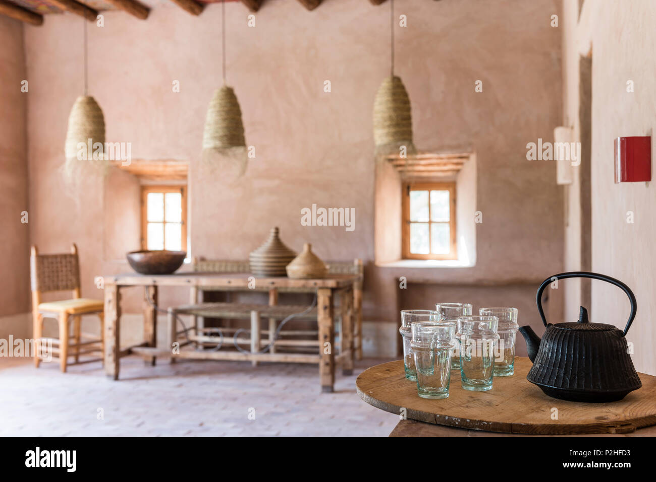 Cast iron tea pot and glasses in foreground of moroccan dining room with rattan oendant lights, earth walls and terracotta floors Stock Photo
