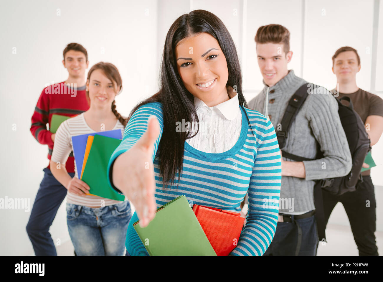 Beautiful smiling girl with books standing in school hall and offering her hand as if to shake hands. Happy group of her friends standing is behind he Stock Photo