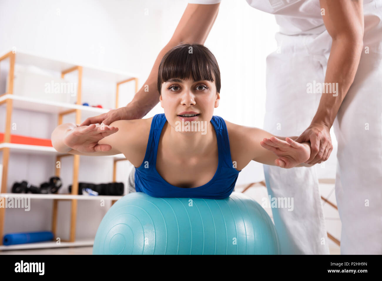 Physiotherapist Assisting Happy Woman While Doing Exercise On Fitness Ball Stock Photo