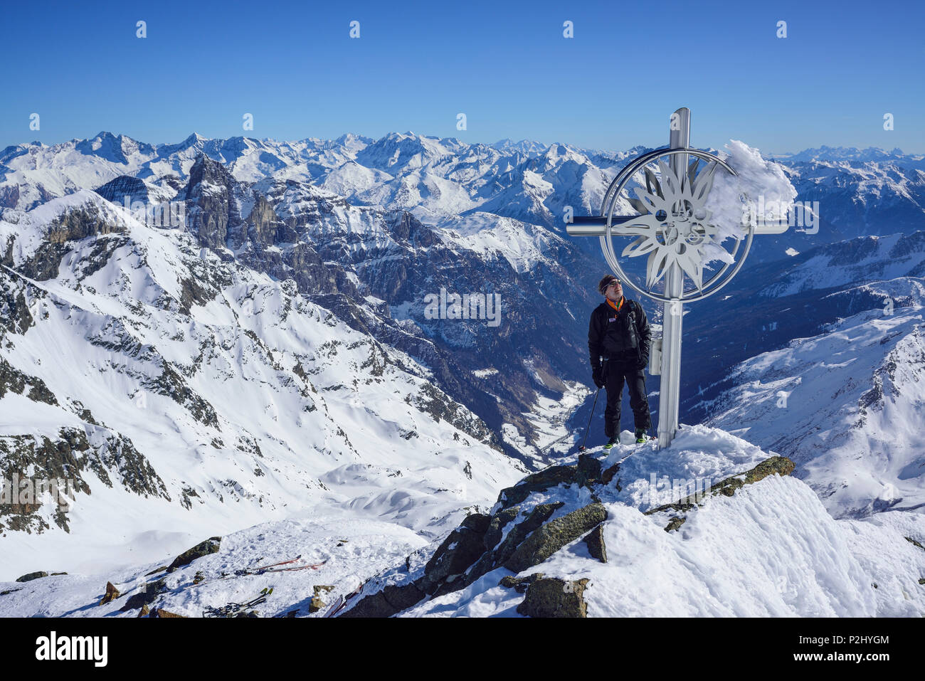 Man back-country skiing standing at cross at summit of Schneespitze, Stubai Alps with Tribulaun and Zillertal Alps in background Stock Photo