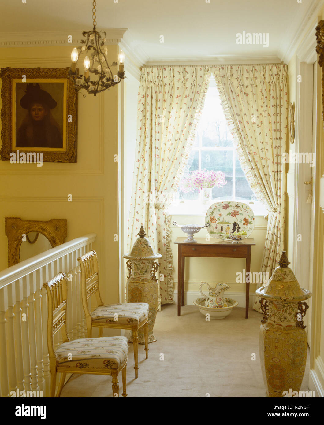 Long cream patterned curtains on window on landing with antique upholstered chairs and tall Chinese vases Stock Photo