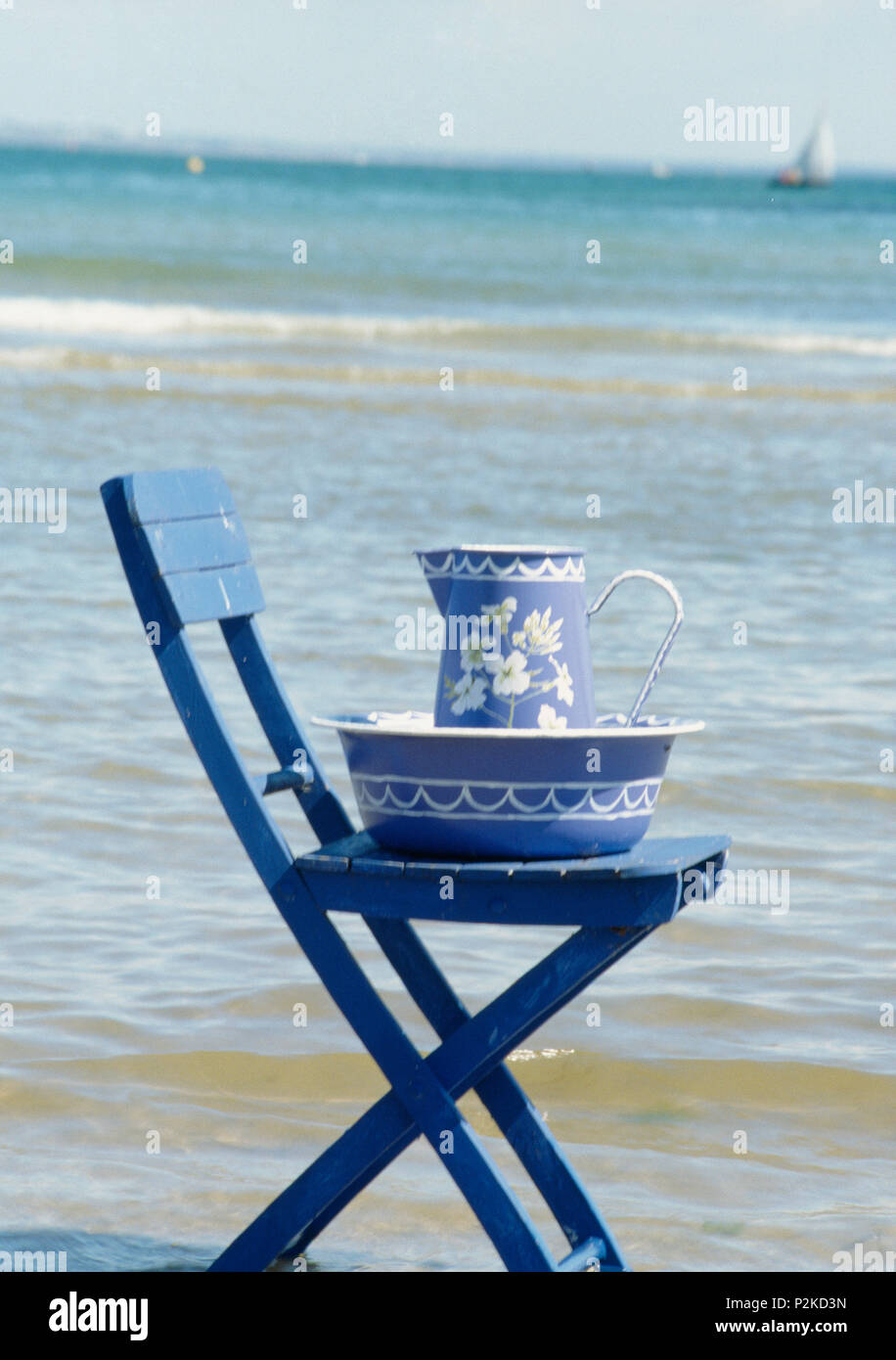 Still-Life of painted blue+white jug and bowl on blue chair beside the sea Stock Photo