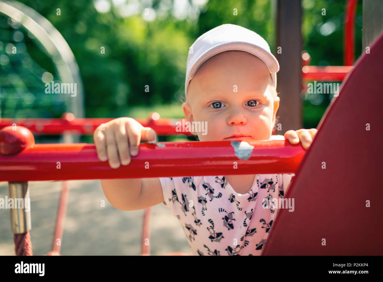 Baby boy playing in playground alone. Portrait of toddler looking at camera with unhappy face. Holding a toy on playground area outdoors. Stock Photo