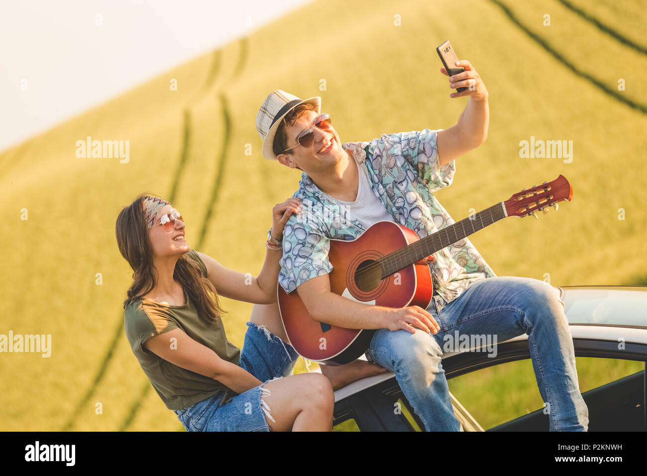 Young hipster couple with acoustic guitar taking selfie while sitting on car roof in nature. Stock Photo