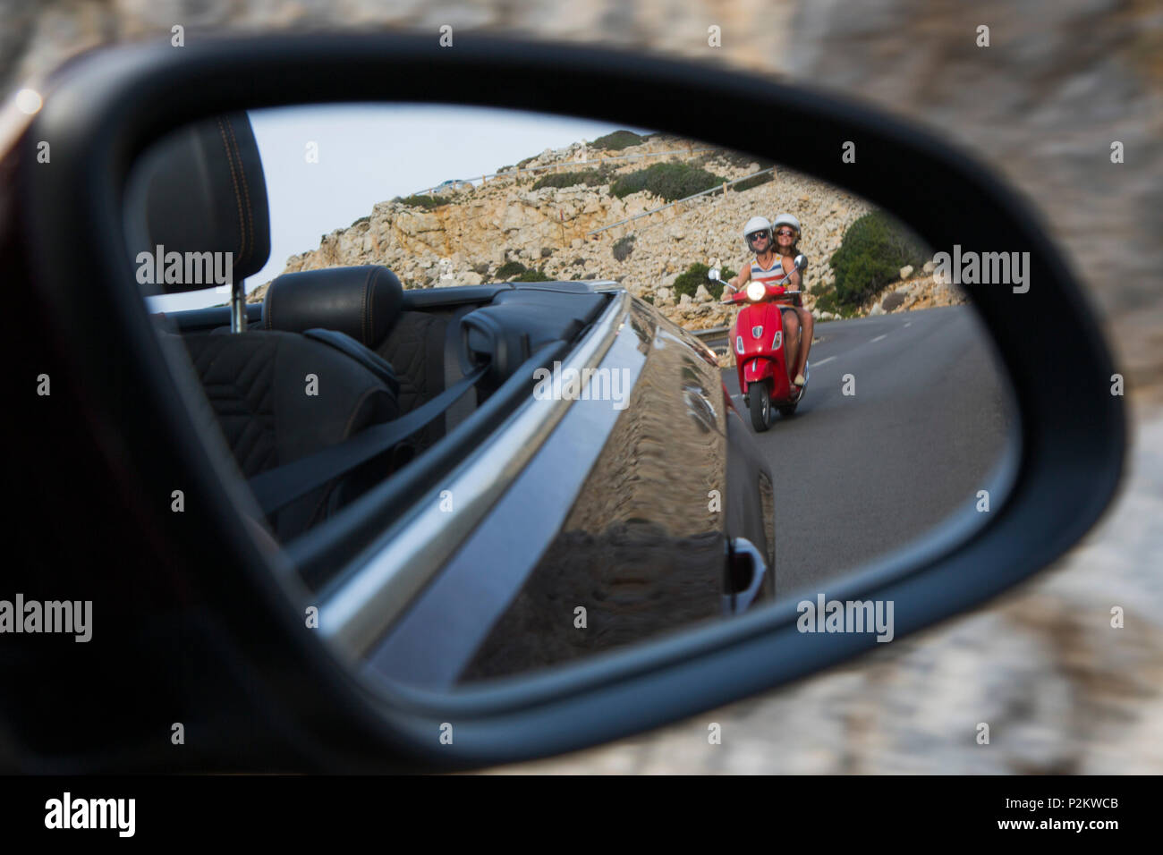 Rear view mirror view of young couple riding a red Vespa scooter on road along Cap de Formentor peninsula with Faro de Formentor Stock Photo