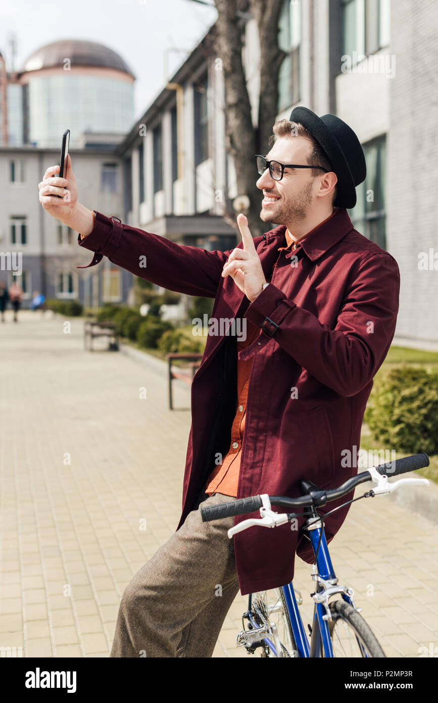 stylish smiling man showing victory sign and taking selfie on smartphone while leaning on bicycle Stock Photo