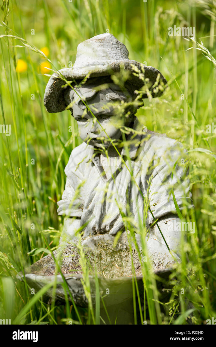 A stone figurine of a boy holding a bird bath in long grass in a garden. Lancashire North West England UK GB Stock Photo