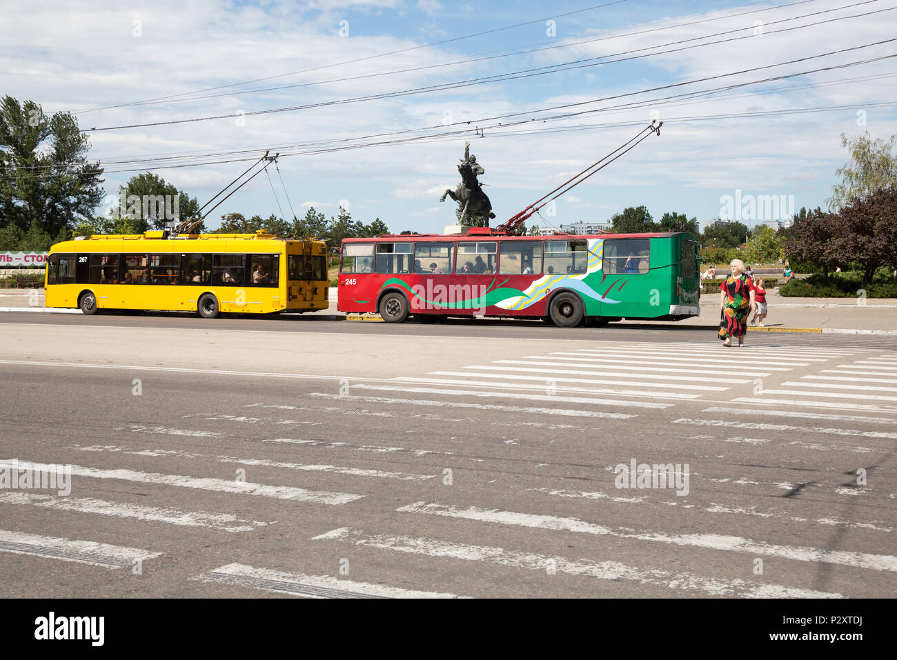 25.08.2016, Tiraspol, Transnistria, Moldova - Trolleybuses, one red-green in the transnistrian colors, older lady in color matching dress crosses Zebr Stock Photo