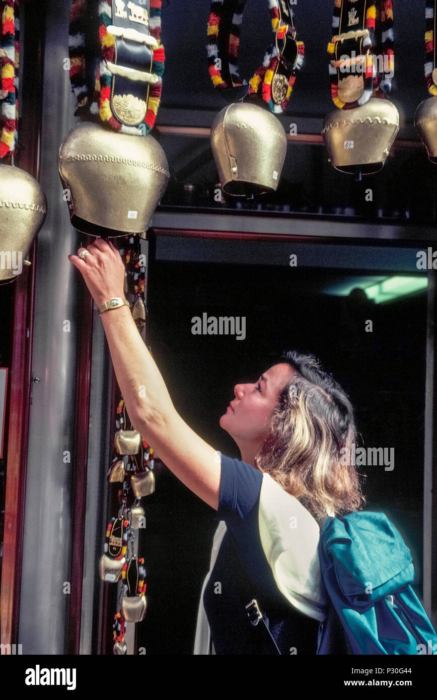 A female traveler inspects large and small Swiss cow bells for sale outside a souvenir shop in Switzerland. An iconic symbol of that European country, the bright metal bells are primarily used by farmers to locate their cattle that graze at large in high alpine meadows during the summer. Noise made by the bells whenever the freely roaming animals move their heads also helps scare off any predators. Stock Photo