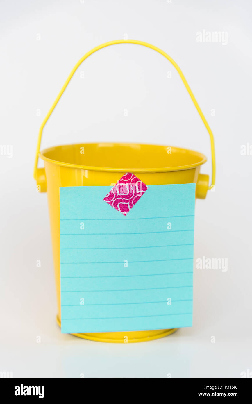 Aqua colored lined paper list taped to front of bright yellow miniature bucket with solid white background to illustrate bucket list Stock Photo