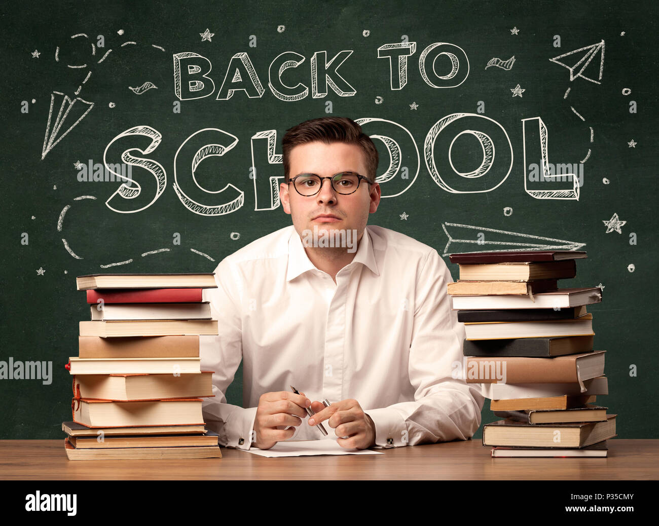 A young teacher in glasses sitting at classroom desk with pile of books ...