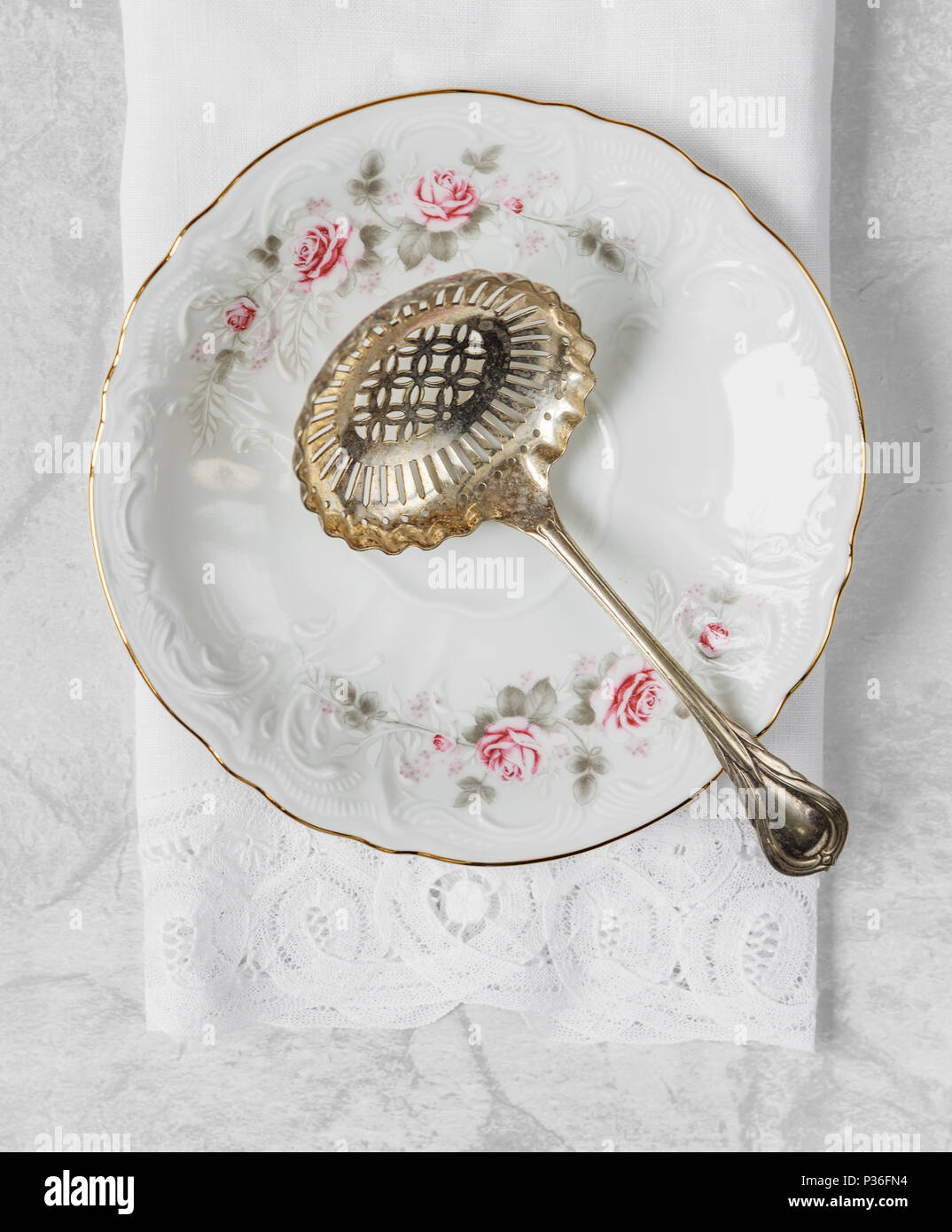 Beautiful silver spoon for compote lies on a porcelain plate with a gold rim, decorated with a pattern of roses, with a lacy linen napkin. Top view Stock Photo