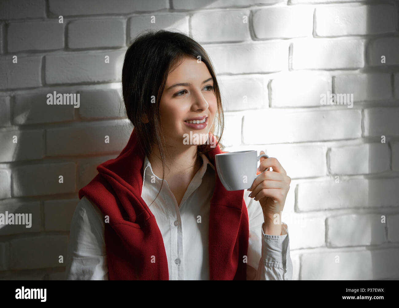 Pretty smiling student drinking coffee in the morning. Having short black hair and light day make up. Wearing casual white shirt and red cardigan. Feeling good, happy. White wall background. Stock Photo