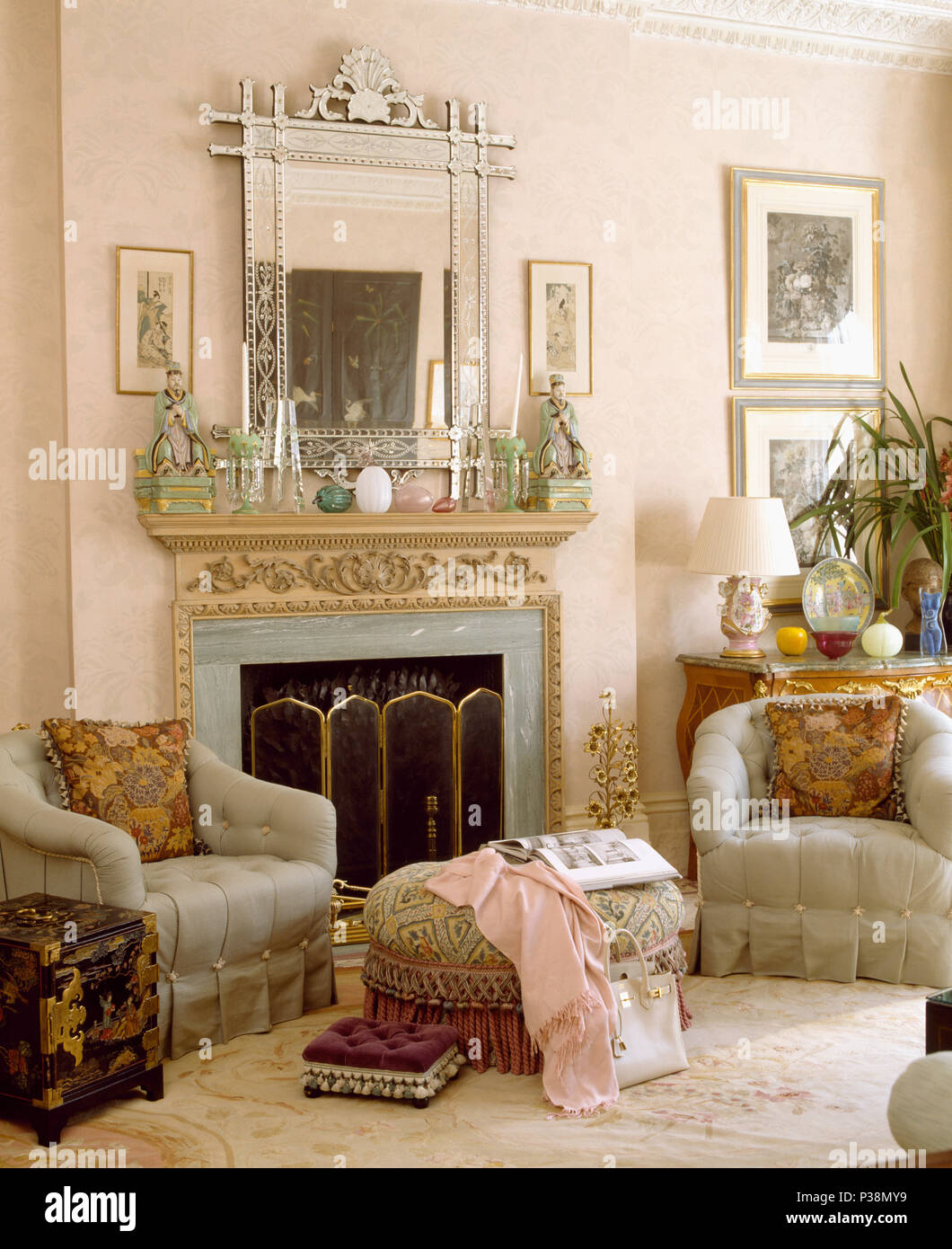 Venetian glass mirror above fireplace with brass firescreen in pale pink living room with grey armchairs Stock Photo