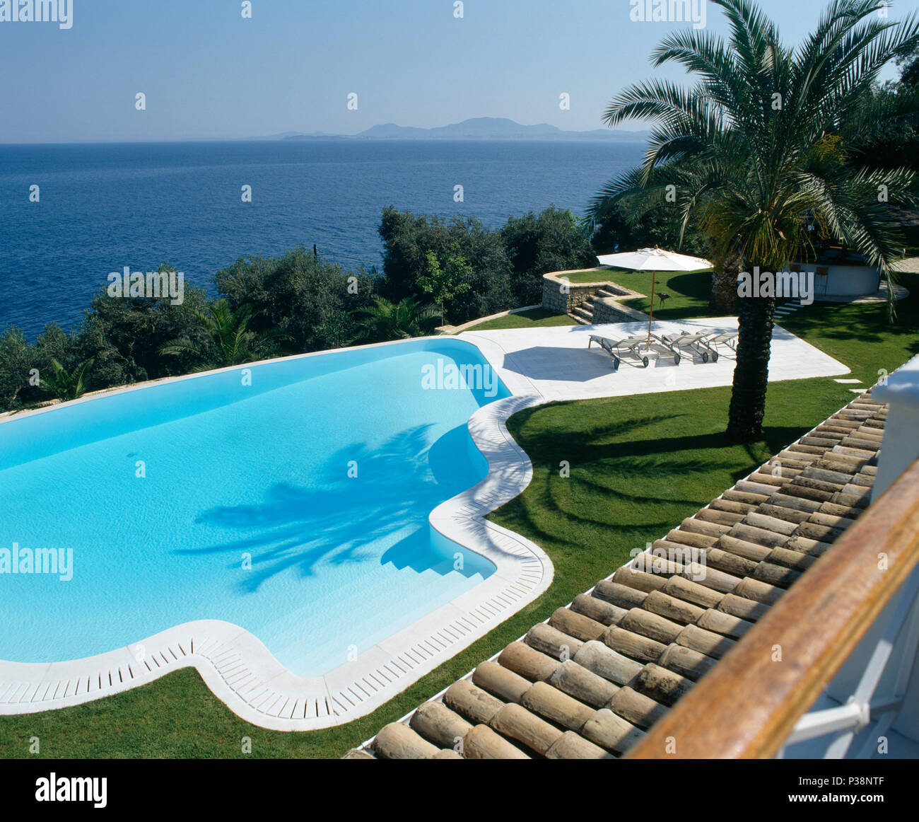 Swimming pool in hillside coastal garden on Corfu with palm tree beside small terrace with view of the ocean Stock Photo