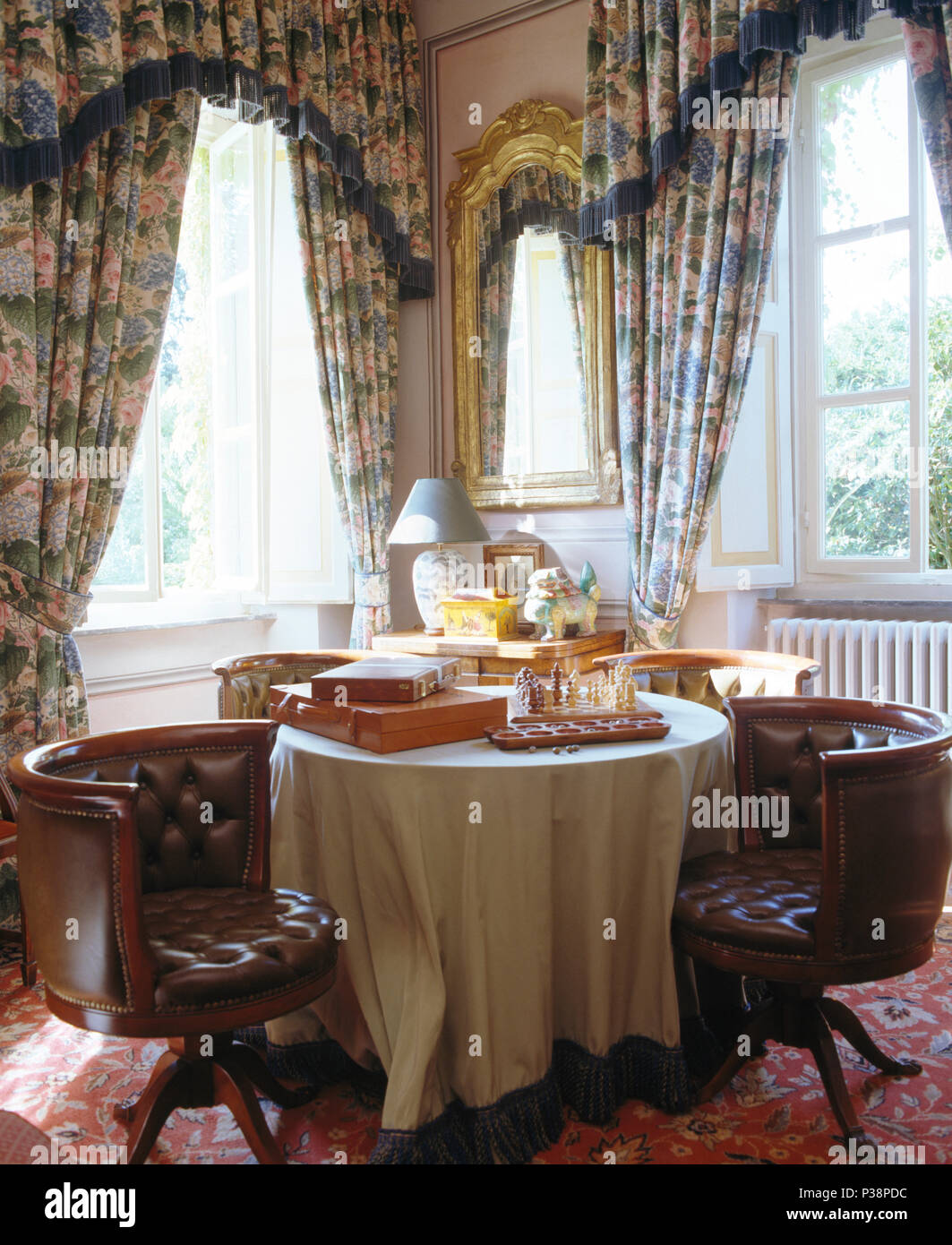 Leather button-back tub chairs at dining table with chess set in front of window with patterned blue curtains Stock Photo