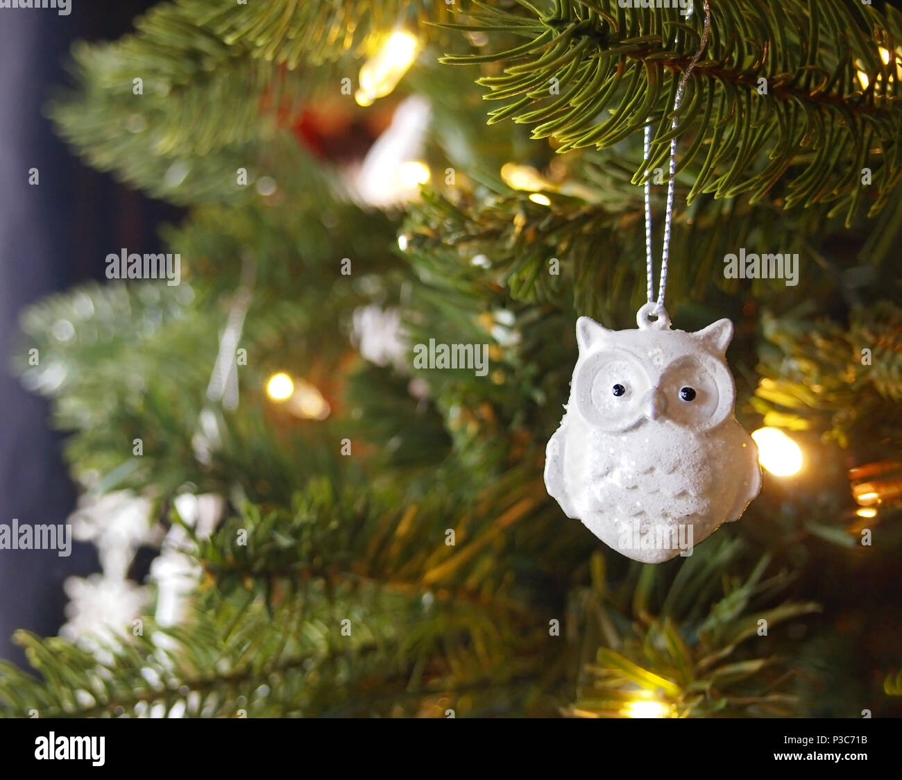 A sparkly white ceramic owl Christmas tree ornament with glitter hangs from a branch of a Christmas tree with glowing golden lights. Stock Photo