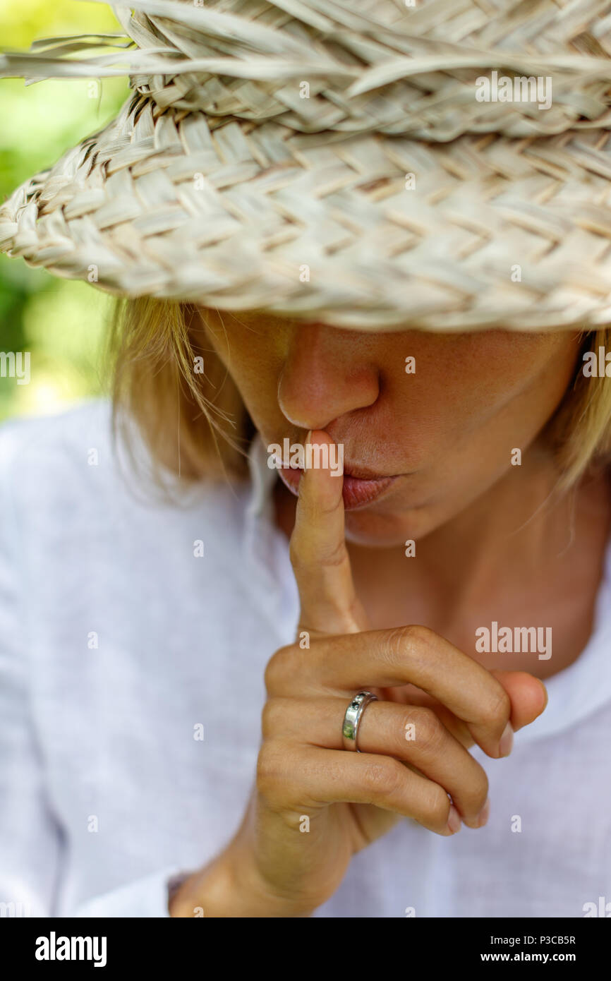 Blond woman in a white shirt and traditional Balinese straw hat with index finger at her lips in silent gesture. Portrait picture. Stock Photo