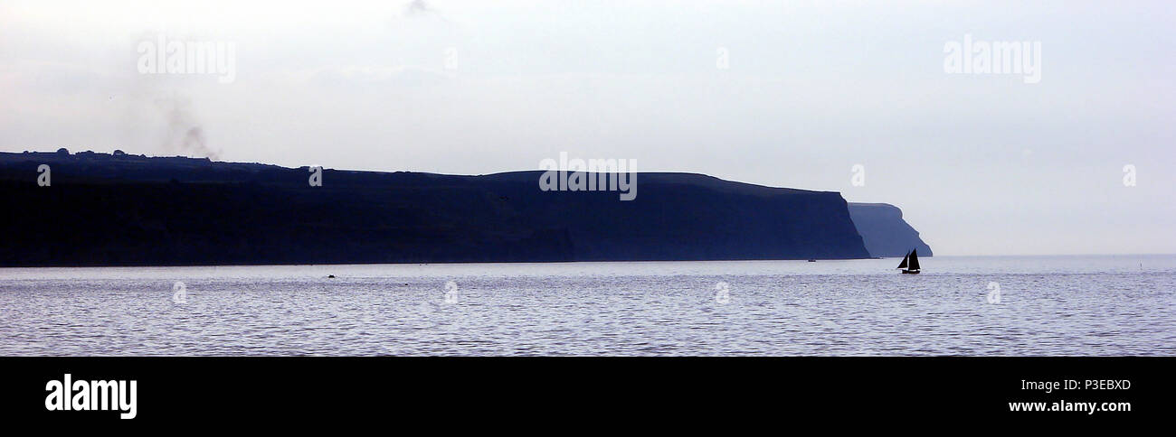 A solitary yachtsman sails into Sandsend Bay, near Whitby, Yorkshire UK at dusk. Kettleness cliffs in background Stock Photo