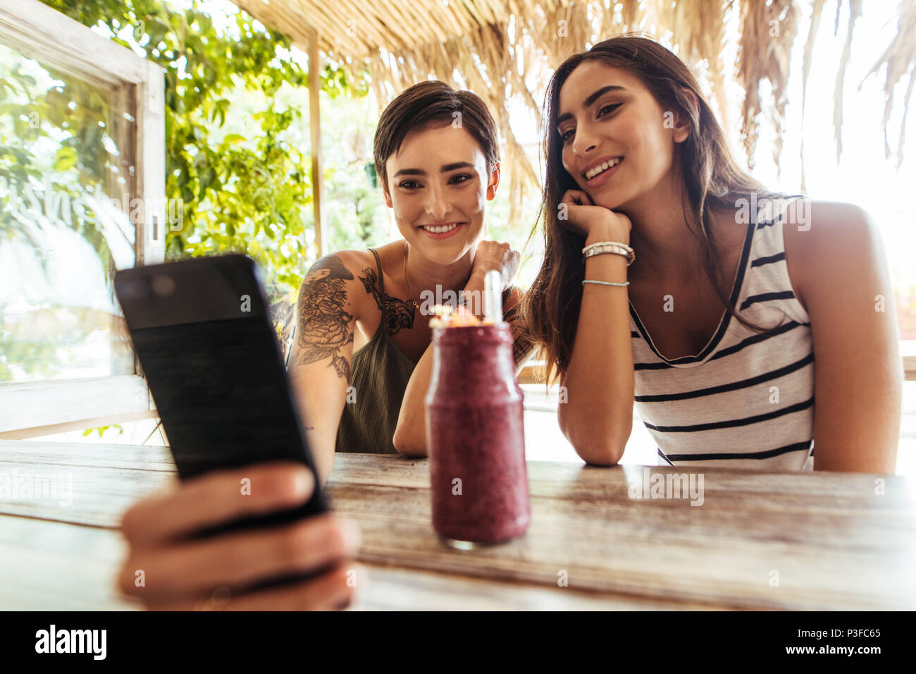 Two smiling women sitting at a restaurant with a smoothie on the table taking photograph. Woman shooting selfie with a friend at a restaurant. Stock Photo