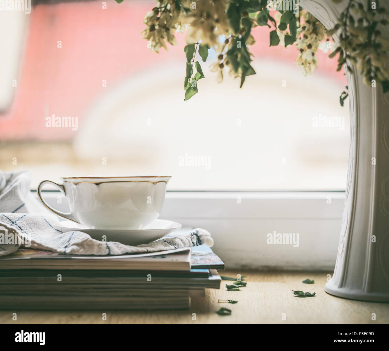 Cup on a stack of magazines at window with vase and flowers. Summer still life. Cozy home scene Stock Photo