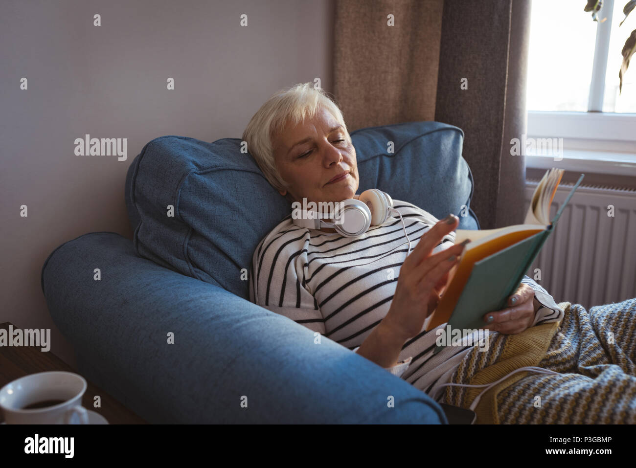Senior woman relaxing on a sofa reading a book in living room Stock Photo