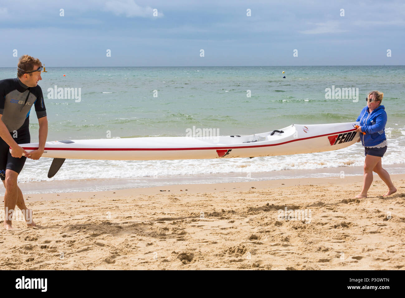 Man and woman carrying a surf ski surfski along seashore at Branksome Chine, Poole, Dorset, England UK in June Stock Photo