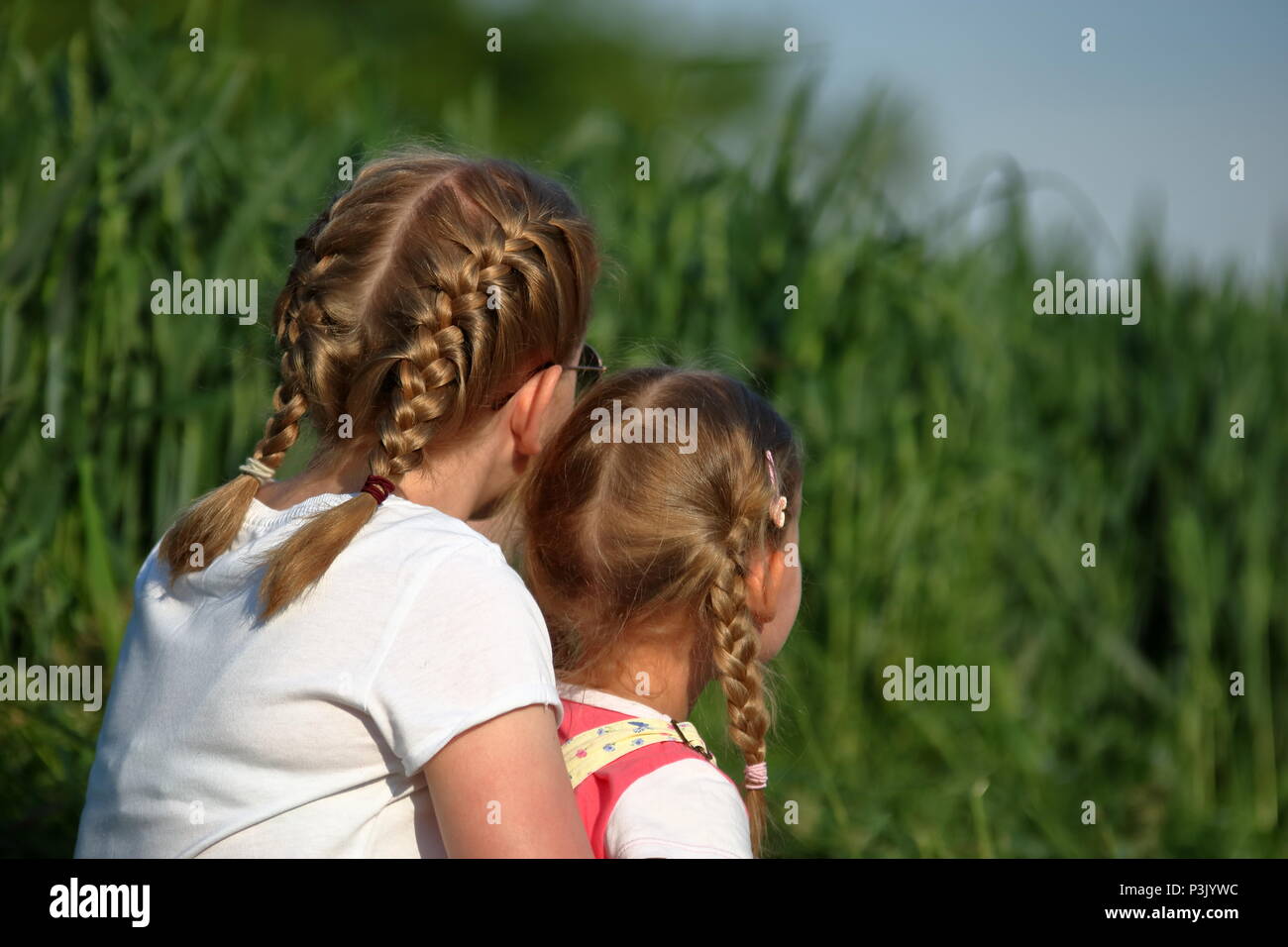 Two blond-haired children, girls, sisters, sit on their back, outdoor, have braids, green plants in background Stock Photo