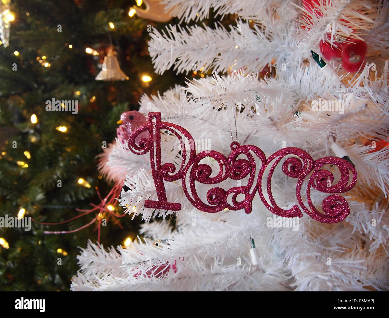 The word PEACE spelled out in pink glittered lettering is an ornament hanging on a white Christmas tree with glowing lighted evergreens in the backgro Stock Photo