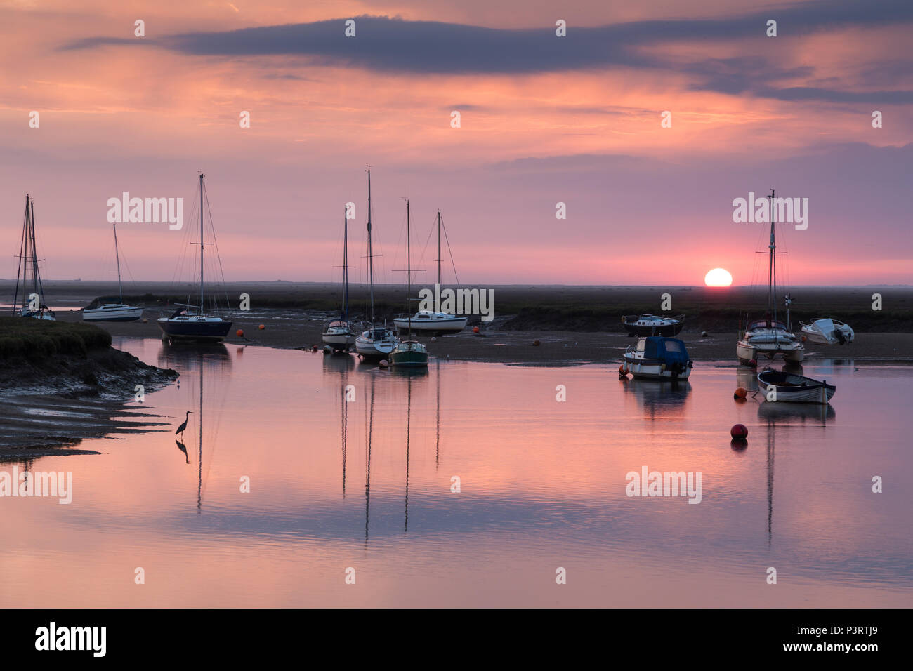 The sun rising behind sailing boats in the harbour at Wells next the Sea with a heron in the foreground. Stock Photo