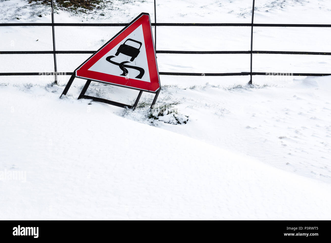 A metal fence partially buried under mounds of drifting snow with a road sign leaning on it indicating slippery roads for vehcile travel. Stock Photo