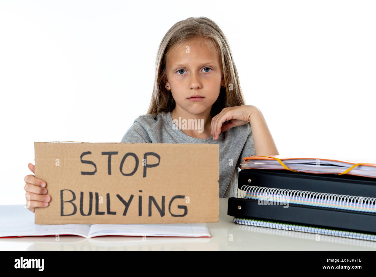 young cute caucasian blonde blue eyed schoolgirl scared and stress holding paper with text stop bullying written looking desperate asking for help sit Stock Photo