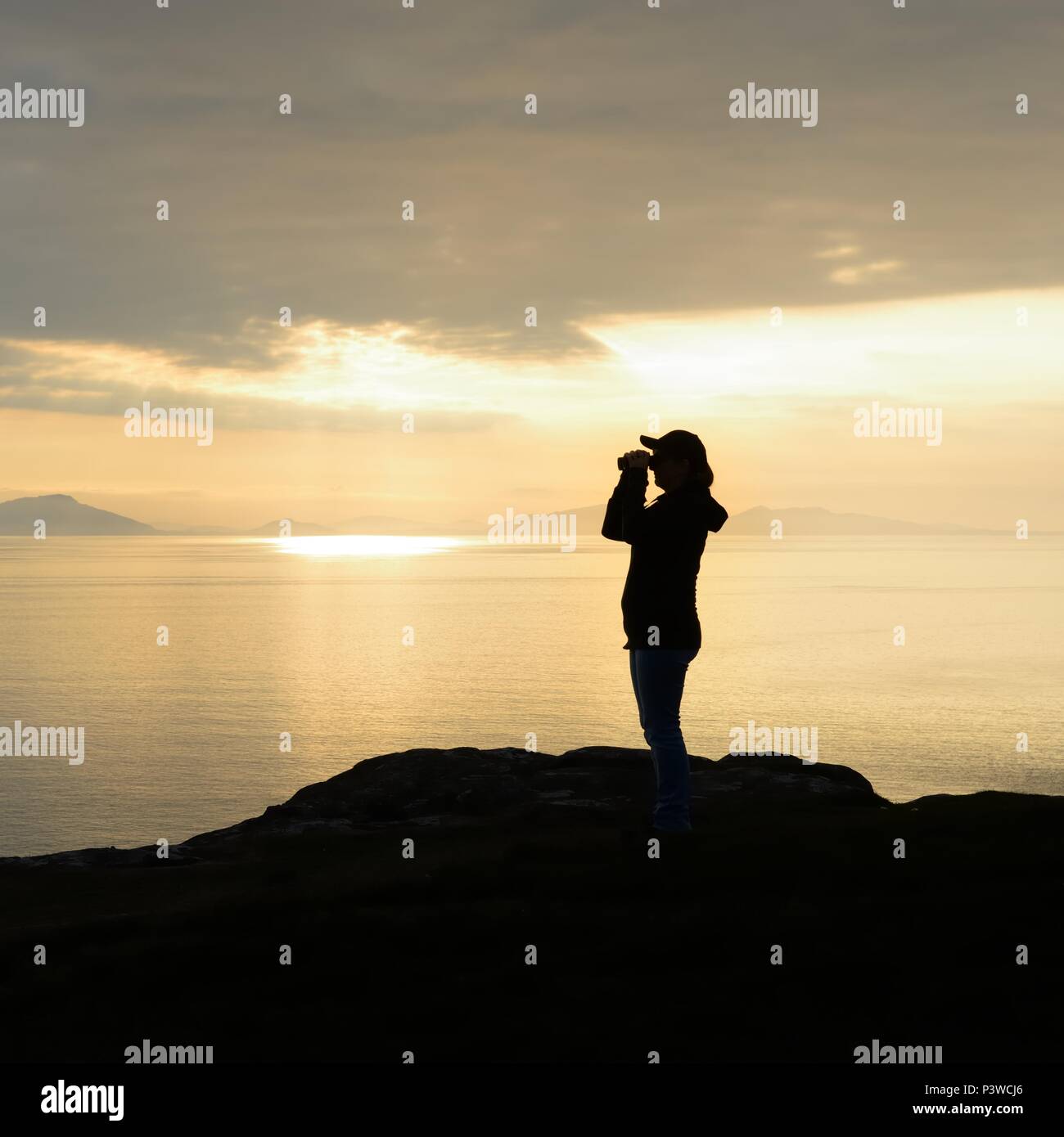 A silhouetted woman using binoculars to view wildlife at sunset along the coastline at Neist Point on the Isle of Skye, Scotland, UK Stock Photo