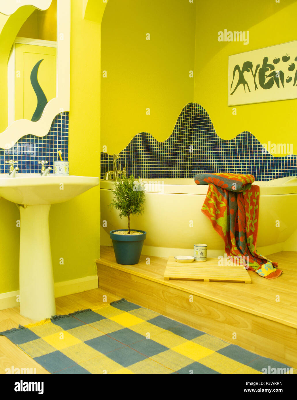 Mosaic tiling above corner bath in yellow split-level bathroom with yellow+blue checked rug below pedestal basin Stock Photo