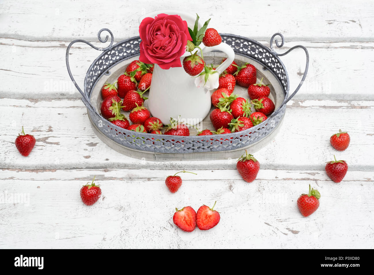 Fresh plump strawberries on white wooden background. A red rose in a white tin can on an old metal tray make a vintage look. Stock Photo