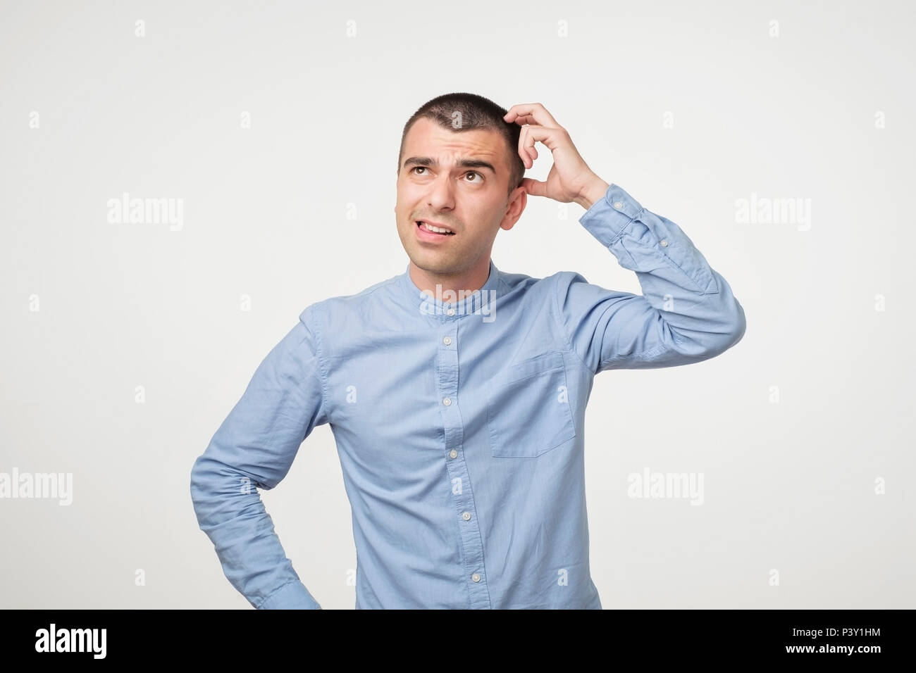 Handsome spanish guy in blue shirt, frowning and looking unsatisfied while scratching head, standing over gray background. He can not remember some fa Stock Photo