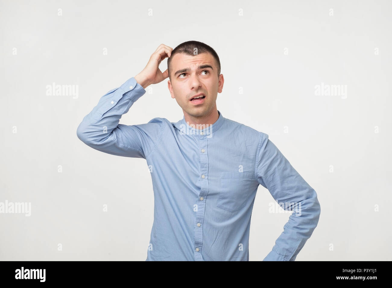 Handsome spanish guy in blue shirt, frowning and looking unsatisfied while scratching head, standing over gray background. He can not remember some fa Stock Photo