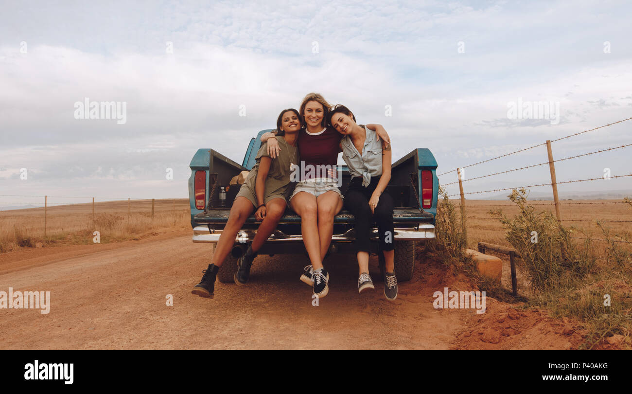 Portrait of three young female friends sitting at the back of a pickup truck parked on a country road. Group of women on a country side road trip. Stock Photo