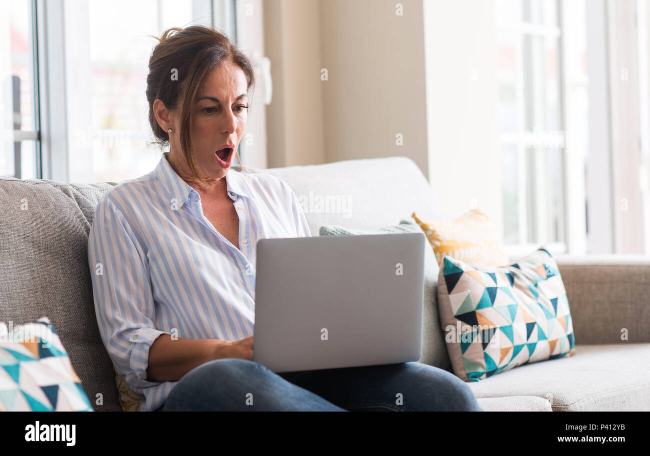 Middle aged woman using laptop in the sofa scared in shock with a surprise face, afraid and excited with fear expression Stock Photo