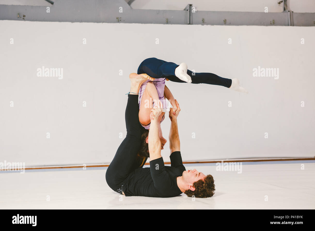 A young Caucasian male and female couple practicing acrobatic yoga in a white gym on mats. They are in the STAR pose. Stock Photo