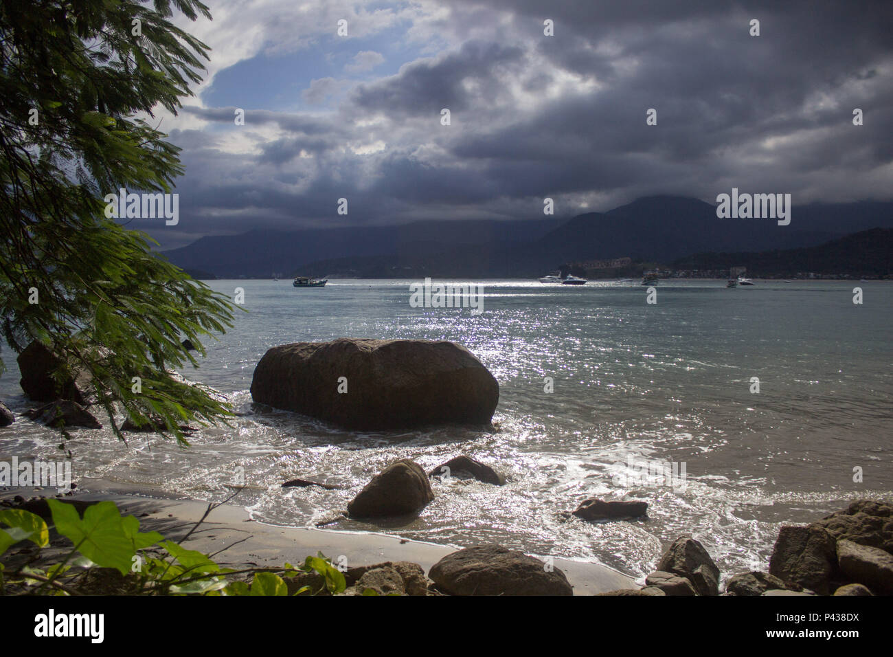 Rochas e pedras em praia ensolarada com barcos navegando no mar ao longe em Ubatuba, SP, Brasil. Stock Photo